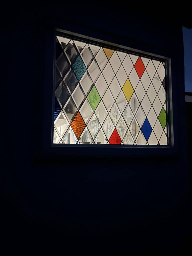 A rectangular stained glass window featuring a harlequin pattern of clear textured diamonds, accented by scattered diamonds in teal, orange, yellow, green, red, and blue, illuminated from within at night.
