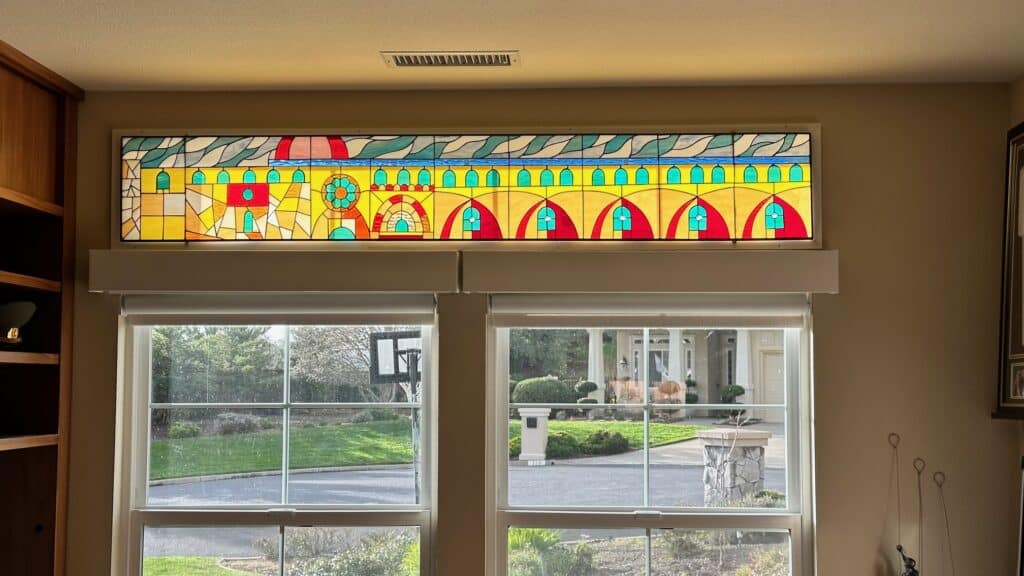 A long, rectangular stained glass transom panel featuring an abstract architectural design with repeating yellow and red arches, teal dome shapes, and a circular medallion, set against a blue sky border, installed above two residential windows.