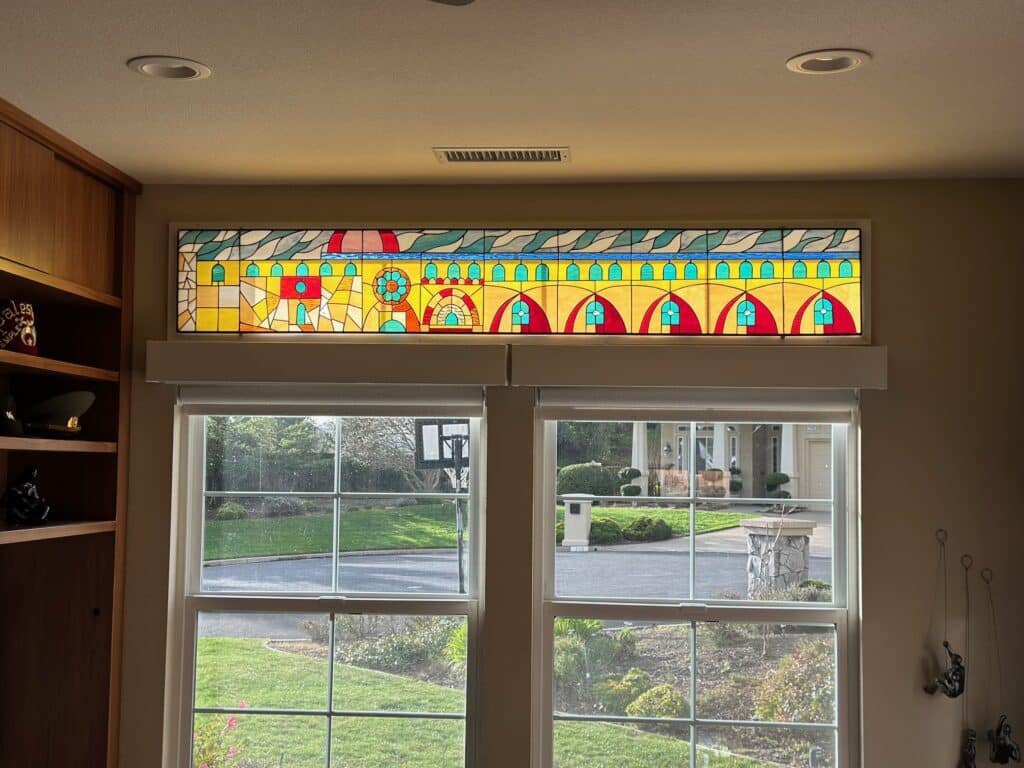A long, rectangular stained glass transom panel with a vibrant, abstract design reminiscent of a stylized cityscape or row of arched buildings, featuring yellow, red, blue, and green geometric and wavy elements, installed above two clear windows.