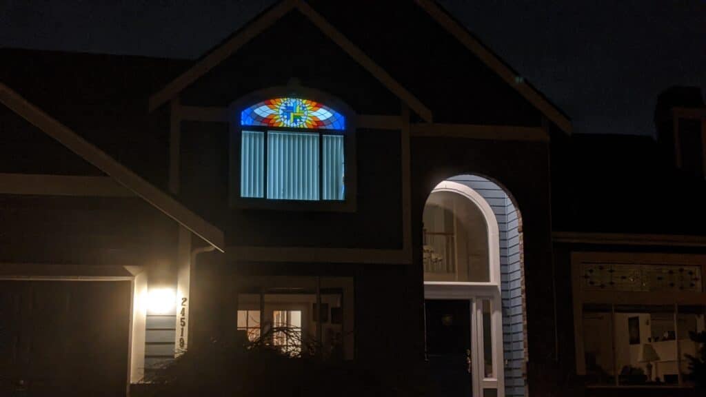A vibrant, arched stained glass window featuring a geometric sunburst design in blue, yellow, orange, and red hues, illuminating a house gable at night.