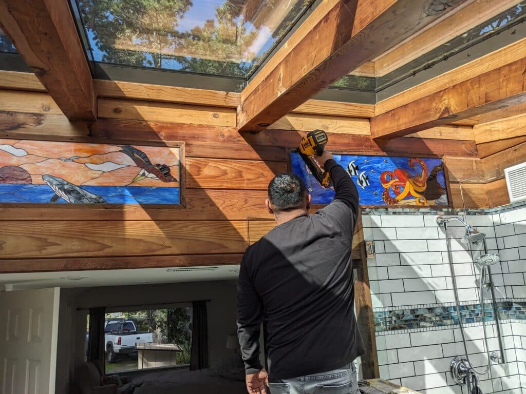 A person installs two rectangular stained glass panels, one depicting a whale and eagle, and the other an octopus and fish, into a rustic wooden wall above a tiled shower area. A skylight above shows trees and blue sky.