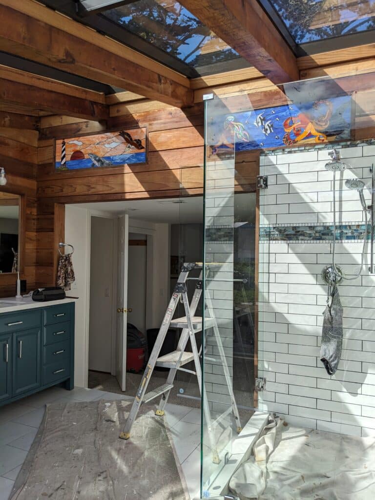 A rustic bathroom with wooden plank walls and ceiling, a glass shower enclosure, and two rectangular stained glass transom panels. The left panel features a lighthouse, a breaching whale, and an eagle over a sunset ocean scene. The right panel displays an orange octopus, a colorful jellyfish, and striped fish in a blue underwater setting. A stepladder and drop cloths indicate ongoing installation.