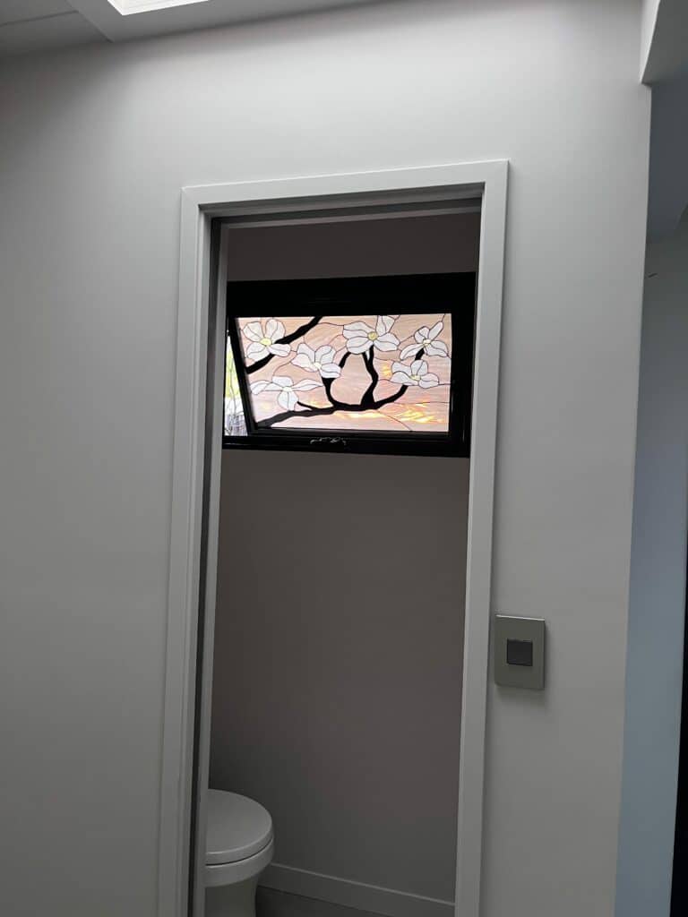 A rectangular stained glass transom window, set in a dark frame, depicting white magnolia blossoms with yellow centers on dark branches, against a textured, light-colored glass background. The window is installed above a doorway.