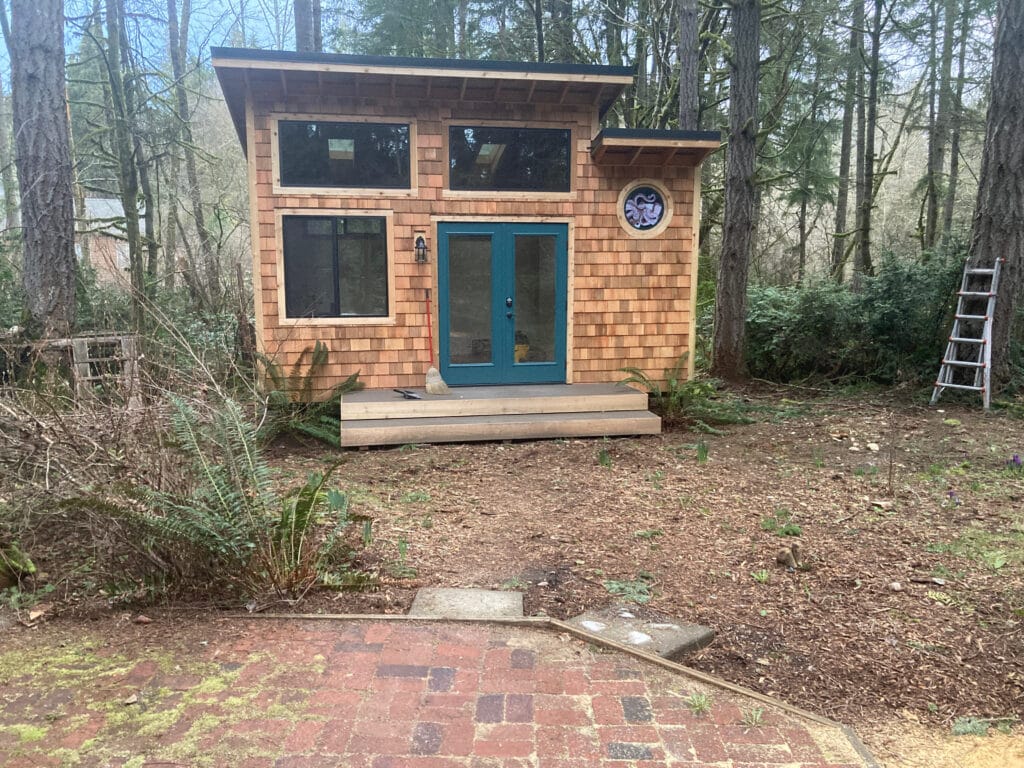 A small, cedar-sided cabin in a forest with teal doors and a circular stained glass window featuring a blue and purple kraken design.