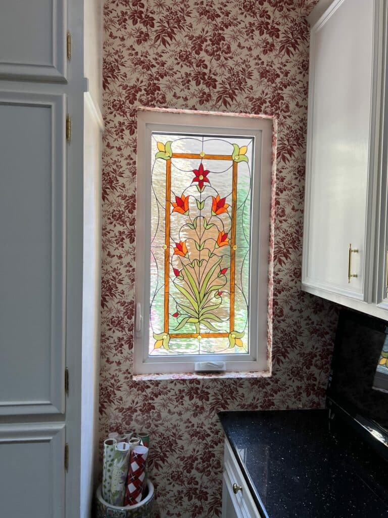 A rectangular stained glass window panel depicting a vibrant floral design with orange and red blossoms, green leaves, and amber accents, set against a clear textured background, encased in a white vinyl window frame. The panel is installed in a room with red and white floral wallpaper, above a black speckled countertop.