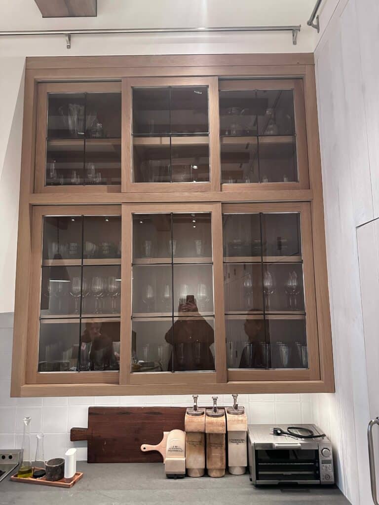 Upper kitchen cabinets with light oak wood frames and clear glass inserts featuring a modern grid pattern, displaying various glassware and dishes. The design allows for open display and light.
