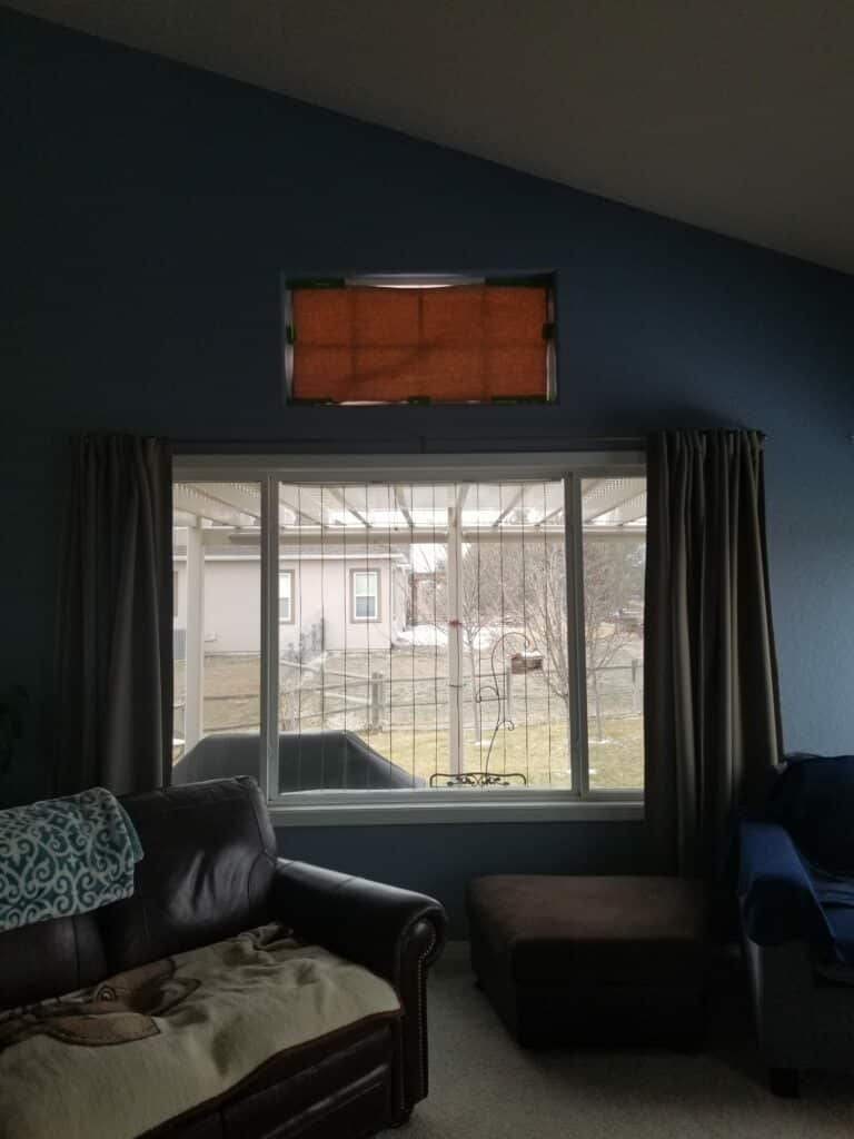 Interior of a living room with dark blue walls, a main horizontal window, and a rectangular transom window above covered by a temporary brown shade. The transom is an ideal location for a custom stained glass panel.