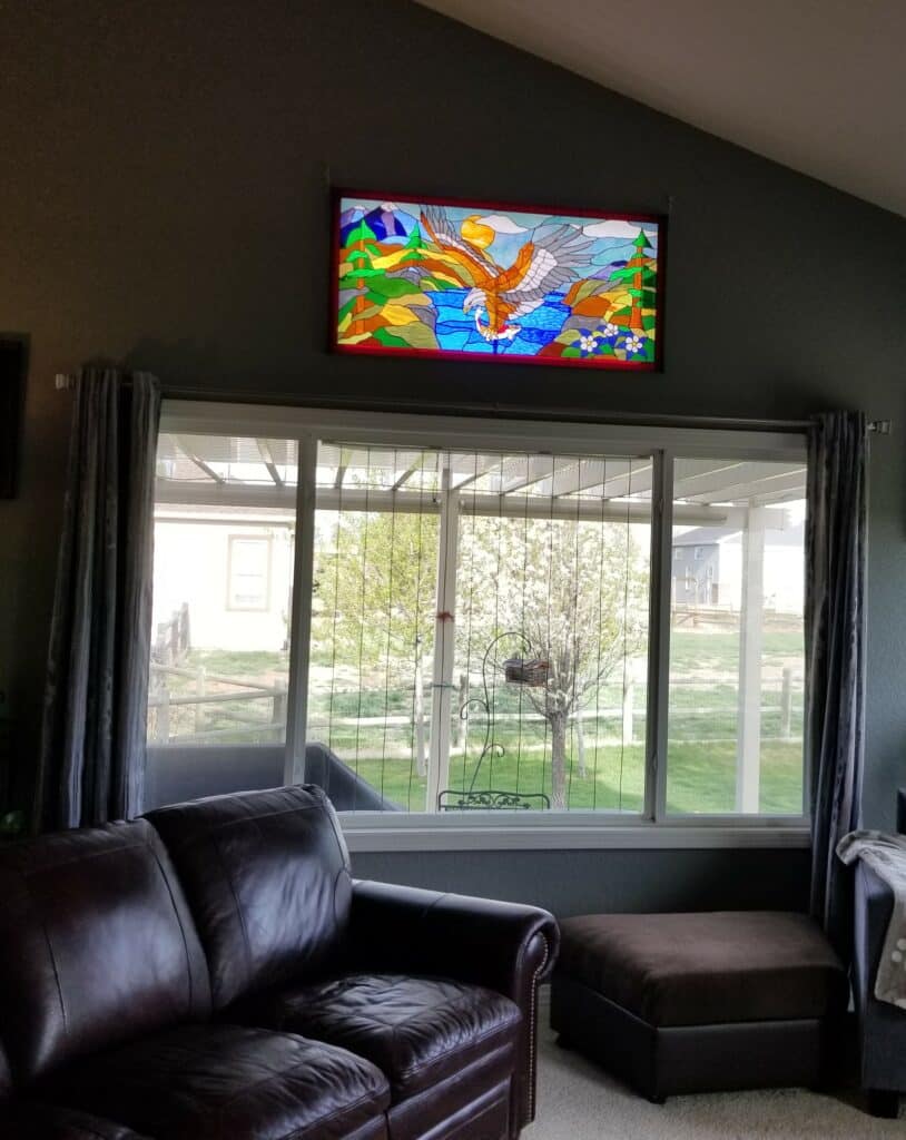 A rectangular stained glass panel depicting a vibrant scene of a golden and white eagle catching a fish from a blue river, with green mountains, pine trees, white flowers, and a yellow sun in the background. The panel is framed in red.