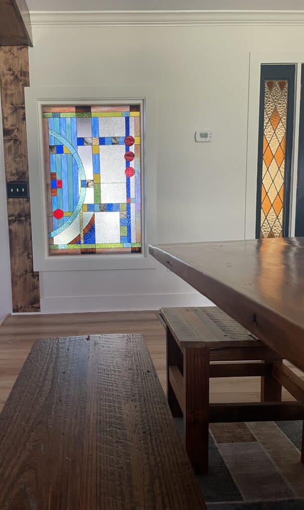 A rectangular stained glass panel with an abstract geometric design featuring various blue and clear textured glass pieces, accented by a large curved light blue element and three red glass circles. A secondary door panel with amber and clear diamond patterned stained glass is visible on the right.