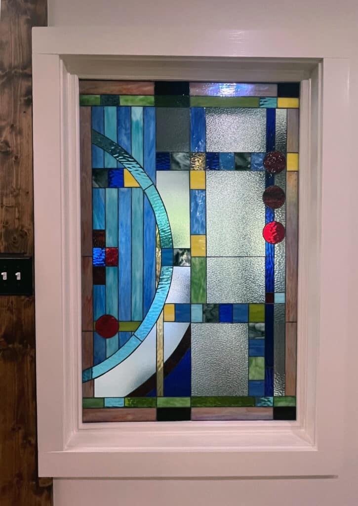 A rectangular stained glass panel in white trim, featuring an abstract geometric pattern with various blues, greens, clear textured glass, and three prominent red circular glass pieces.