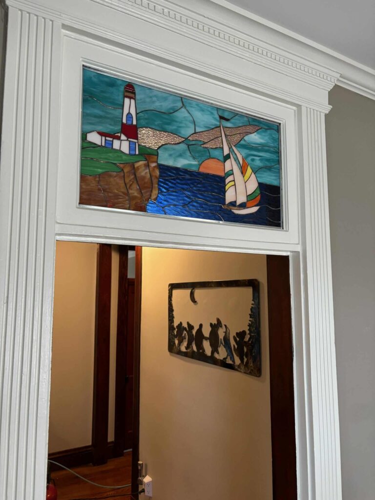 A rectangular stained glass transom depicting a lighthouse on a brown cliff overlooking a blue ocean with a sailboat and an orange sunset.