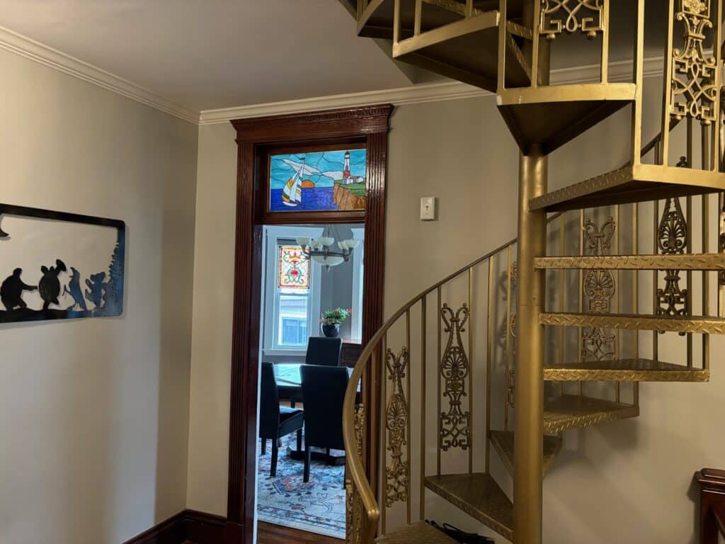 A rectangular stained glass transom depicting a white lighthouse on a brown cliff, a sailboat with colorful sails on a blue sea, and an orange sunset, framed in dark wood above a doorway. A second geometric stained glass panel is visible through the doorway.