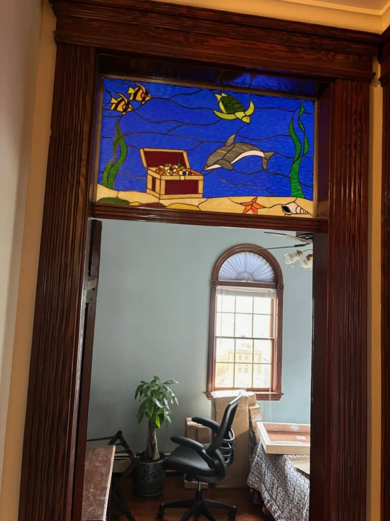 A rectangular stained glass transom panel depicting a vibrant underwater scene with a dolphin, sea turtle, two yellow fish, an open treasure chest filled with gold, seaweed, a red starfish, and a white shell, all against a blue water background and sandy seafloor, installed in an ornate dark wooden doorway.