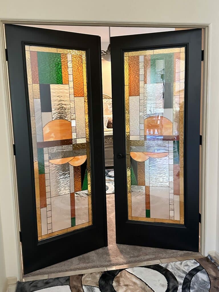 A pair of black French doors with abstract geometric stained glass panels, featuring textured clear, amber, and green art glass, including prominent amber semi-circular elements.