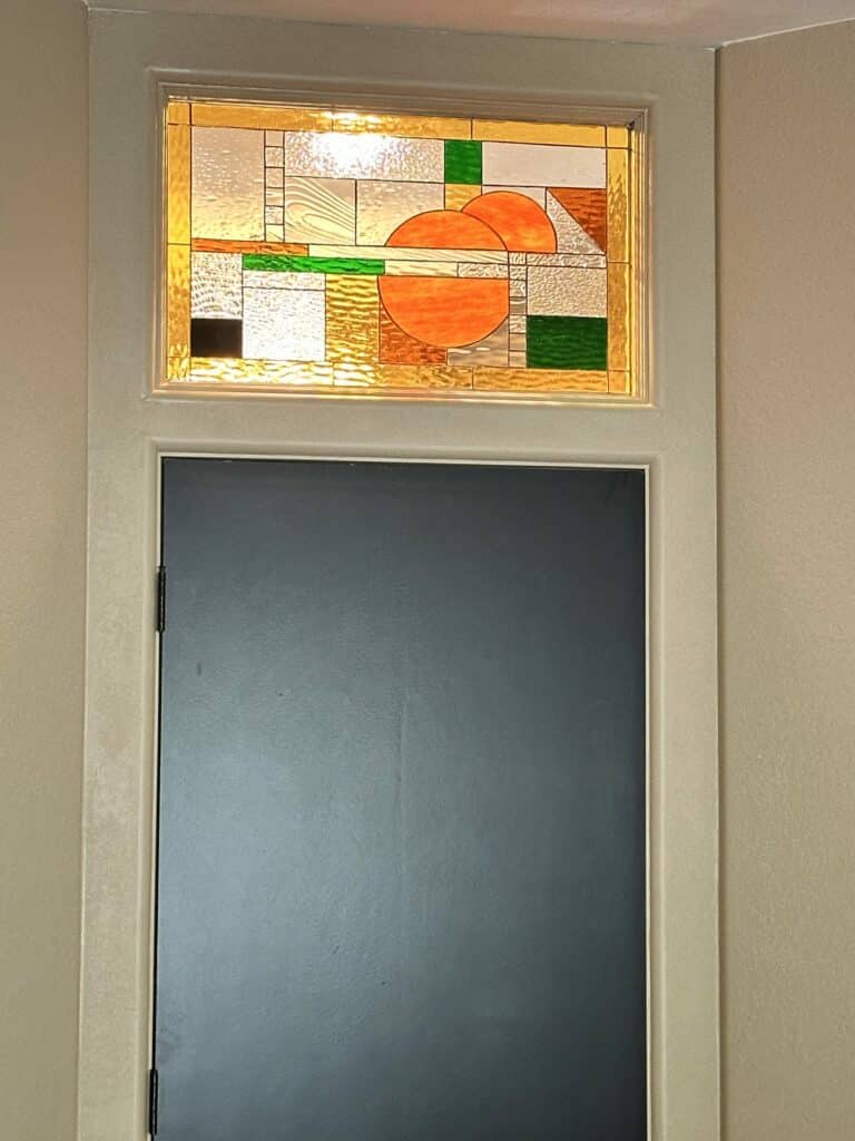 A rectangular leaded stained glass transom window with an abstract geometric design featuring three prominent orange semicircles, green and amber rectangular blocks, and various clear textured glass panels, installed above a dark interior door.