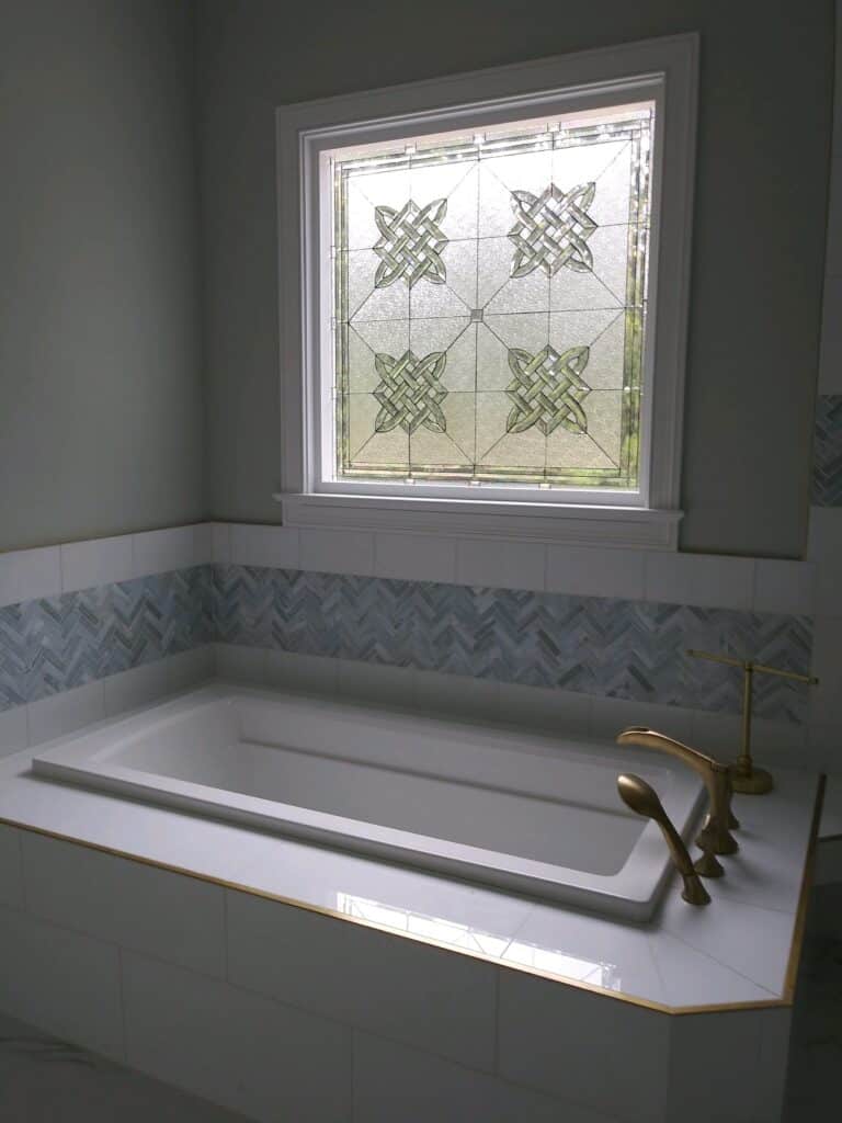 A rectangular clear textured stained glass window featuring a repeating geometric Celtic knot pattern, installed above a built-in white bathtub with brass fixtures and herringbone tile accent.