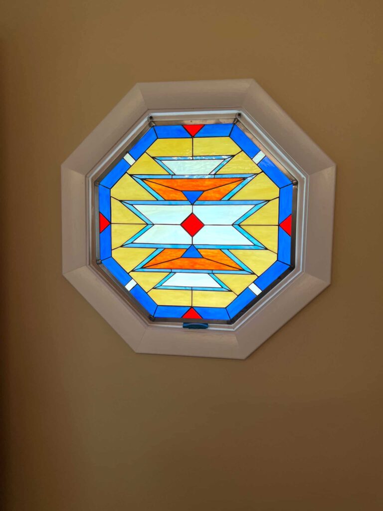 An octagonal stained glass window featuring a vibrant geometric pattern resembling a Navajo rug design, with horizontal zigzag bands in blue, orange, white, and yellow glass, set in a white frame against a light brown wall.