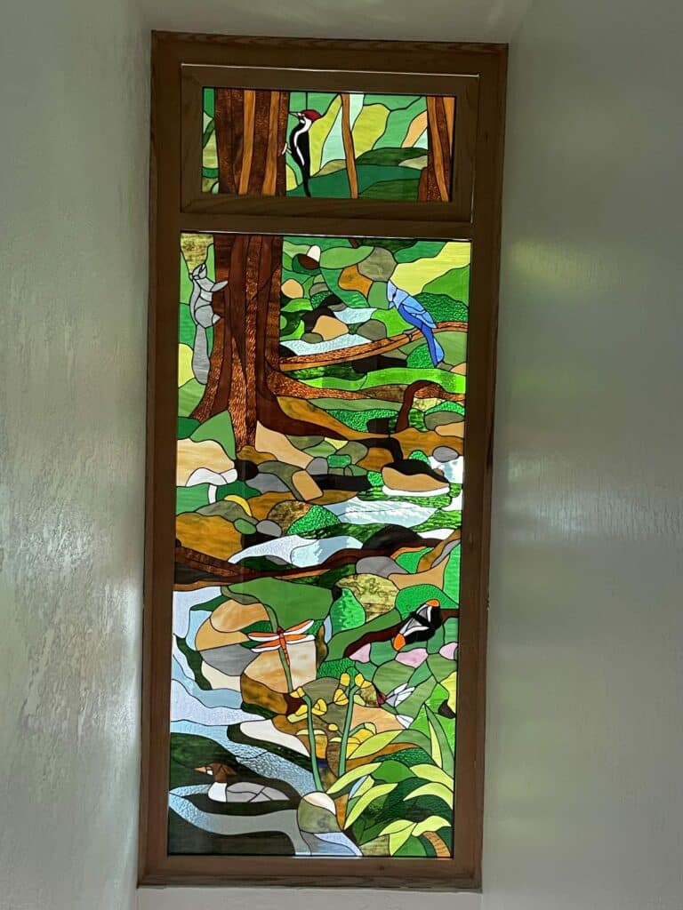 A tall, rectangular stained glass window featuring a vibrant woodland and river scene with various animals. The upper section shows a woodpecker on a tree. The main panel depicts a gray squirrel on a tree, a blue jay on a branch, a colorful bird, an orange dragonfly, and a duck in a flowing stream, surrounded by green foliage, brown trees, and natural elements.