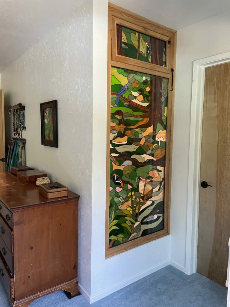 A tall, vertical stained glass panel integrated into a wall, depicting a vibrant woodland scene with a woodpecker, blue jay, butterfly, dragonfly, mushrooms, and trees, all framed in natural wood.