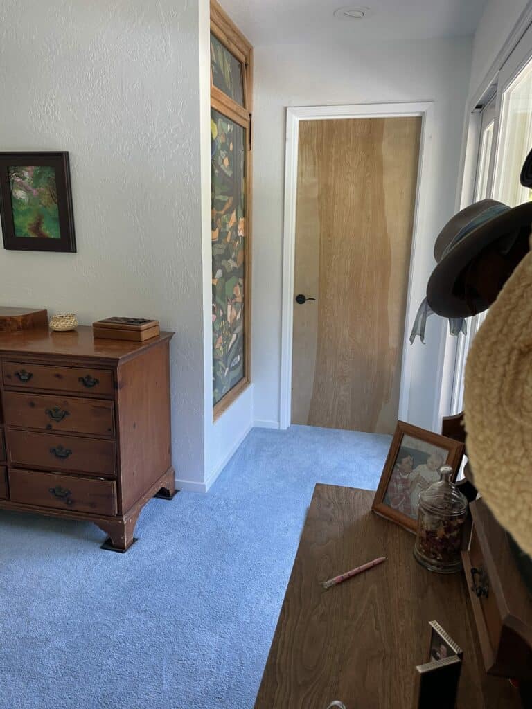 A tall, narrow, vertically installed stained glass panel featuring an abstract organic design with muted green, orange, and brown shapes, set in a natural wood frame.