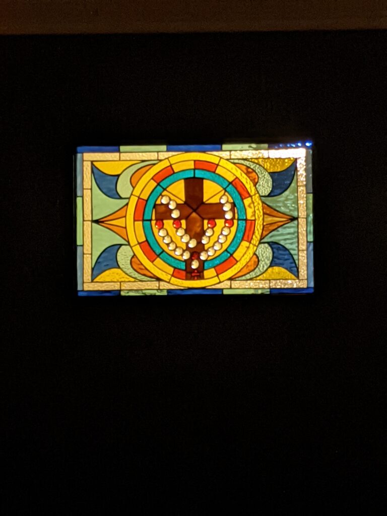 A rectangular stained glass panel featuring a central circular medallion with a brown cross and a draped rosary of white and red beads, surrounded by colorful geometric patterns.