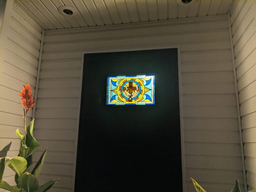 A rectangular, illuminated stained glass panel with a central beaded cross design and geometric patterns in blue, yellow, and orange, set into a dark door on a white paneled wall.