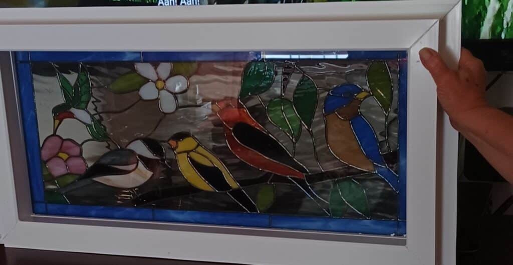 A rectangular stained glass panel in a white frame, depicting a vibrant garden scene with multiple birds and flowers. From left to right, a red-headed hummingbird hovers near pink blossoms, a black-capped chickadee perches below it, followed by a yellow goldfinch and a red cardinal. On the far right, a bluebird is perched, all surrounded by green leaves and white flowers, with a deep blue border framing the main artwork.