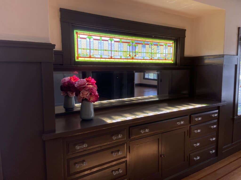 A horizontal, leaded stained glass transom window featuring a geometric Prairie style design with green, blue, and amber glass. It is set above a dark stained wooden built-in buffet with a mirrored backsplash. A vase of pink and red flowers sits on the buffet.