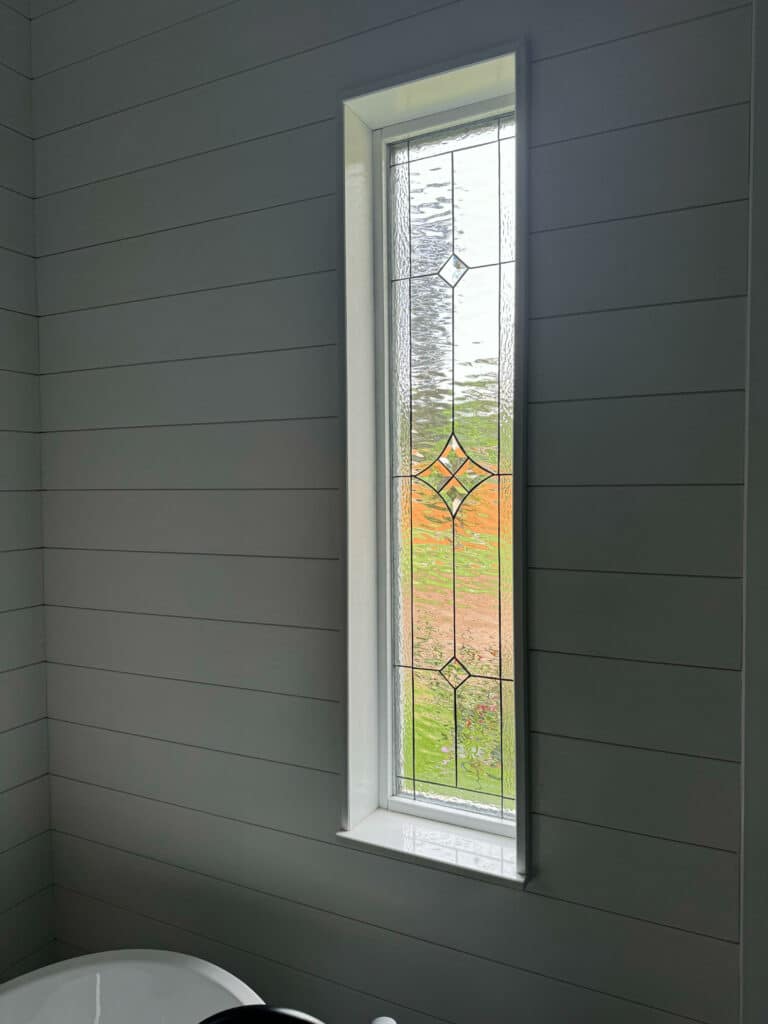 A tall, narrow clear stained glass window featuring textured glass and geometric bevels, set in a light gray shiplap wall.