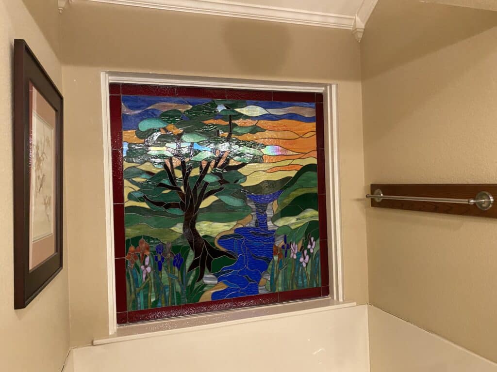 A rectangular stained glass panel depicting a vibrant landscape with a large tree, a blue river and waterfall, green hills, and a sunset sky, bordered in red.