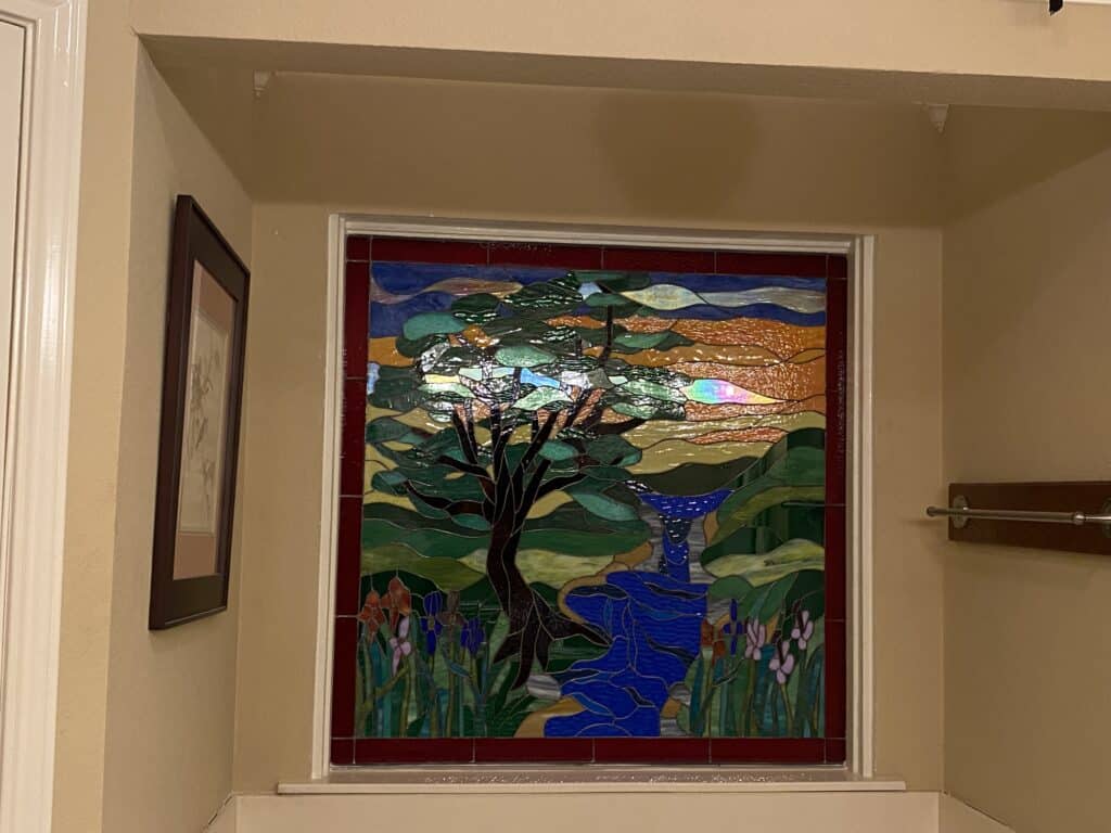 A rectangular leaded stained glass panel depicting a vibrant landscape with a prominent dark tree, a blue river flowing through hills, colorful flowers, and a sunset sky with orange, yellow, and blue hues.