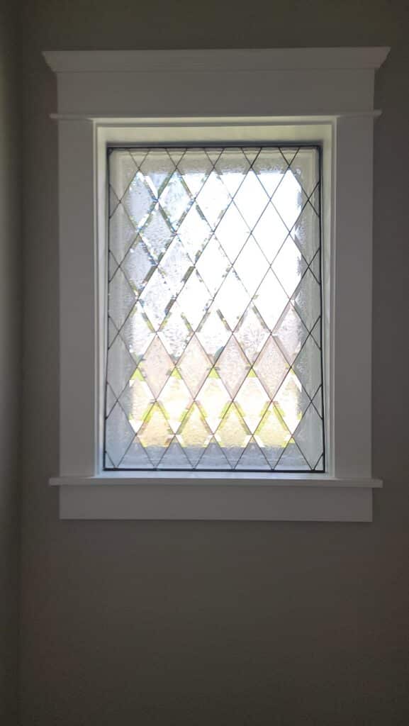 A rectangular clear leaded stained glass window featuring a uniform pattern of diamond-shaped textured panes, framed in white against a gray wall.