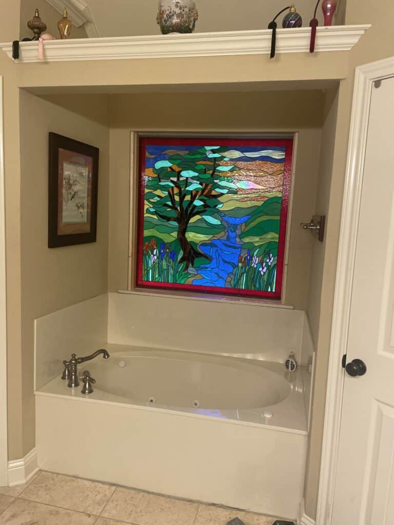 A rectangular stained glass window in a bathroom alcove, featuring a vibrant landscape with a large tree, a winding blue river, green hills, and purple and white irises, installed above a white corner bathtub.