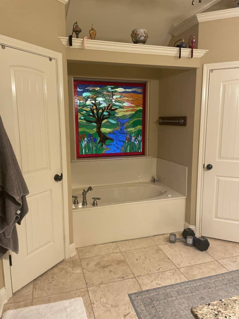 A rectangular stained glass window above a white bathtub, featuring a nature scene with a large brown tree, a blue river with a waterfall, green hills, purple and orange irises, and a multi-colored sky, all bordered in red.