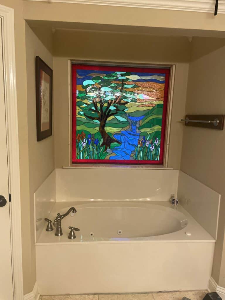 A rectangular stained glass window depicting a vibrant nature scene with a large brown tree, a blue winding river, green hills, and colorful irises, framed in red and installed above a white jetted bathtub.