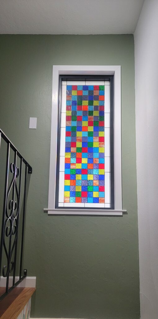 A rectangular stained glass panel featuring a vibrant checkerboard pattern of colorful, textured glass squares, framed in white.