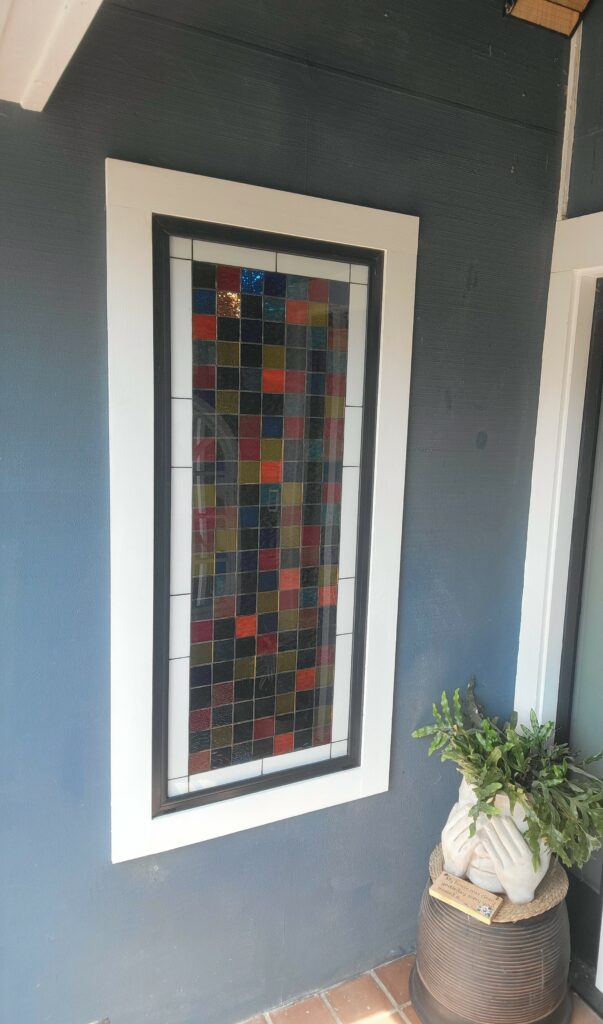 A rectangular stained glass panel installed in a dark blue wall, featuring a grid of multi-colored and clear textured square glass pieces, framed by black and white trim.