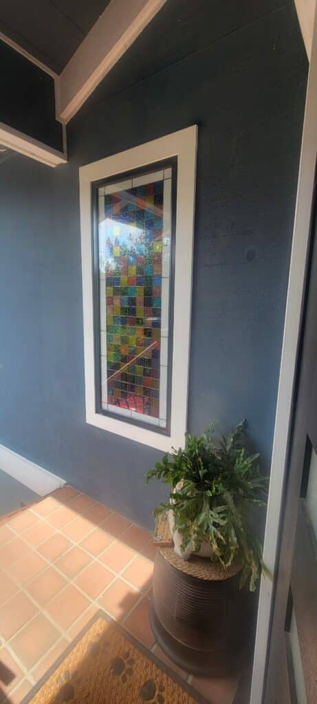 A vertical rectangular stained glass panel featuring a mosaic of small, textured, colorful square glass pieces, providing privacy in an entryway. It is set in a white frame on a dark blue wall with a potted plant below.