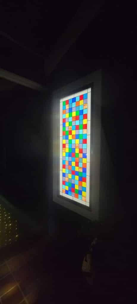 A rectangular stained glass panel featuring a multicolored grid of small squares in red, blue, green, and yellow, glowing brightly in a dark room.