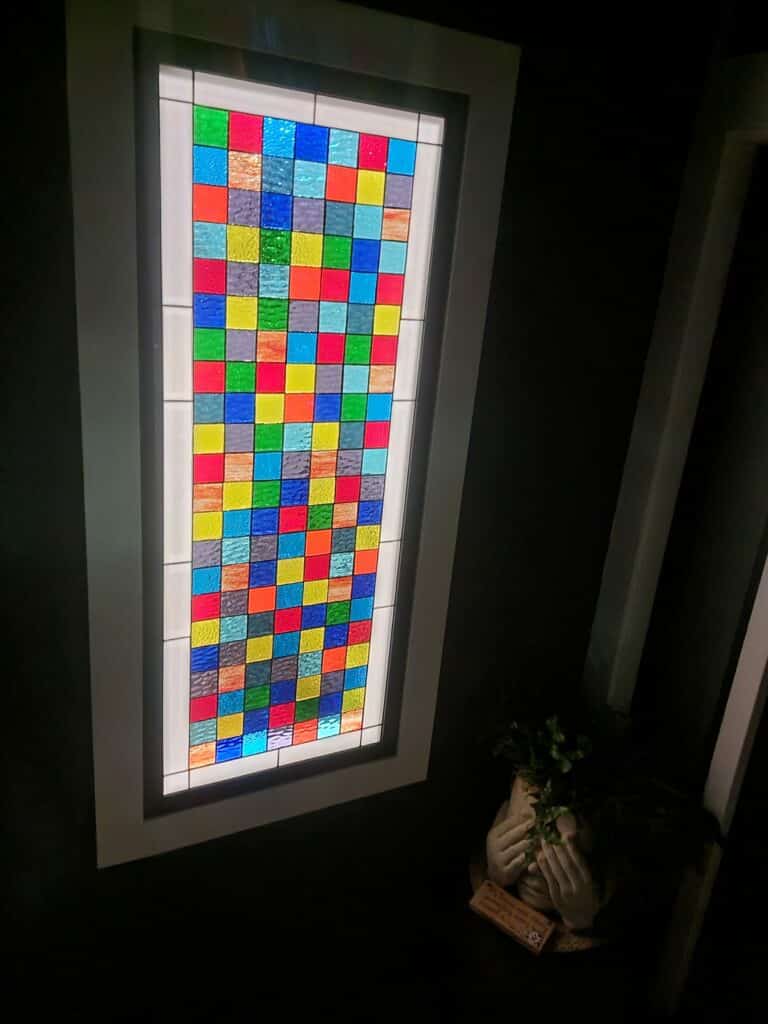 A tall, rectangular stained glass window panel featuring a vibrant checkerboard pattern of small, textured square glass pieces in red, blue, green, yellow, orange, and light blue, set within a clear glass border and white frame.