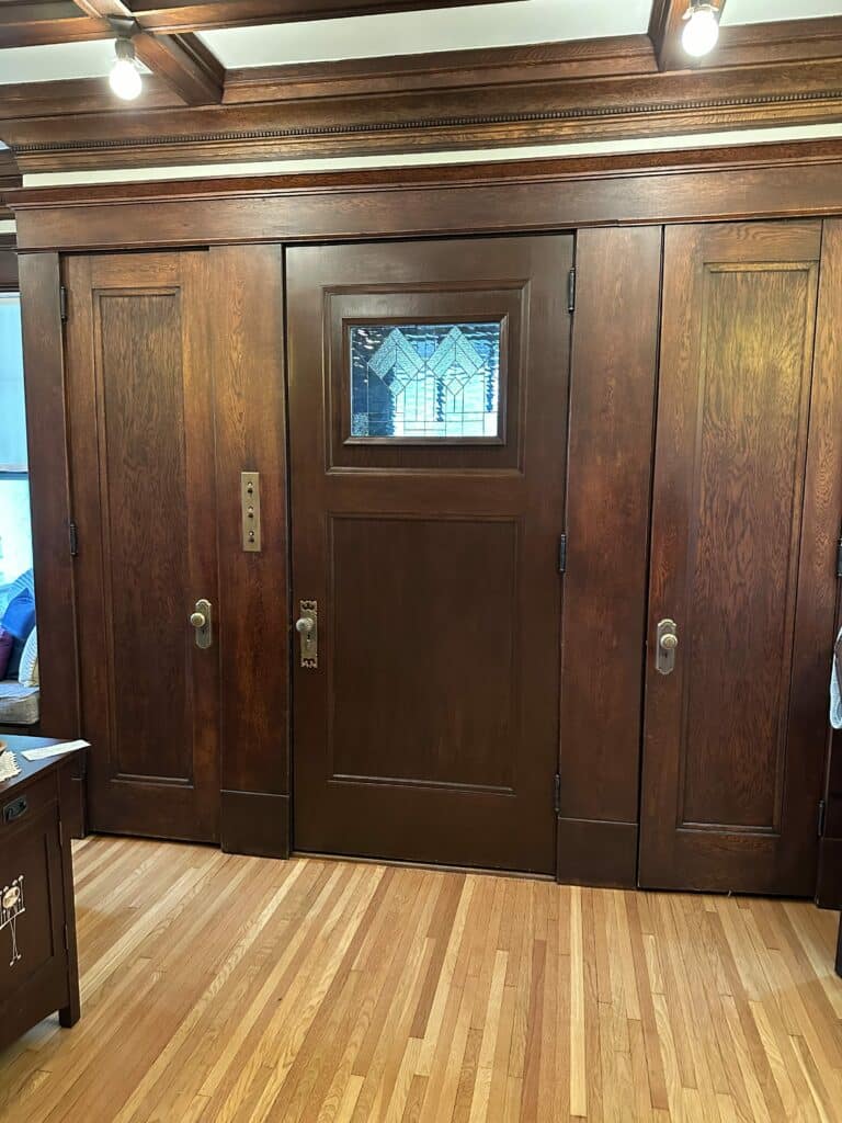 A dark wood panel door featuring a rectangular stained glass insert with clear textured and light blue geometric patterns, flanked by tall wooden doors or panels, set within a rich wood-paneled interior.