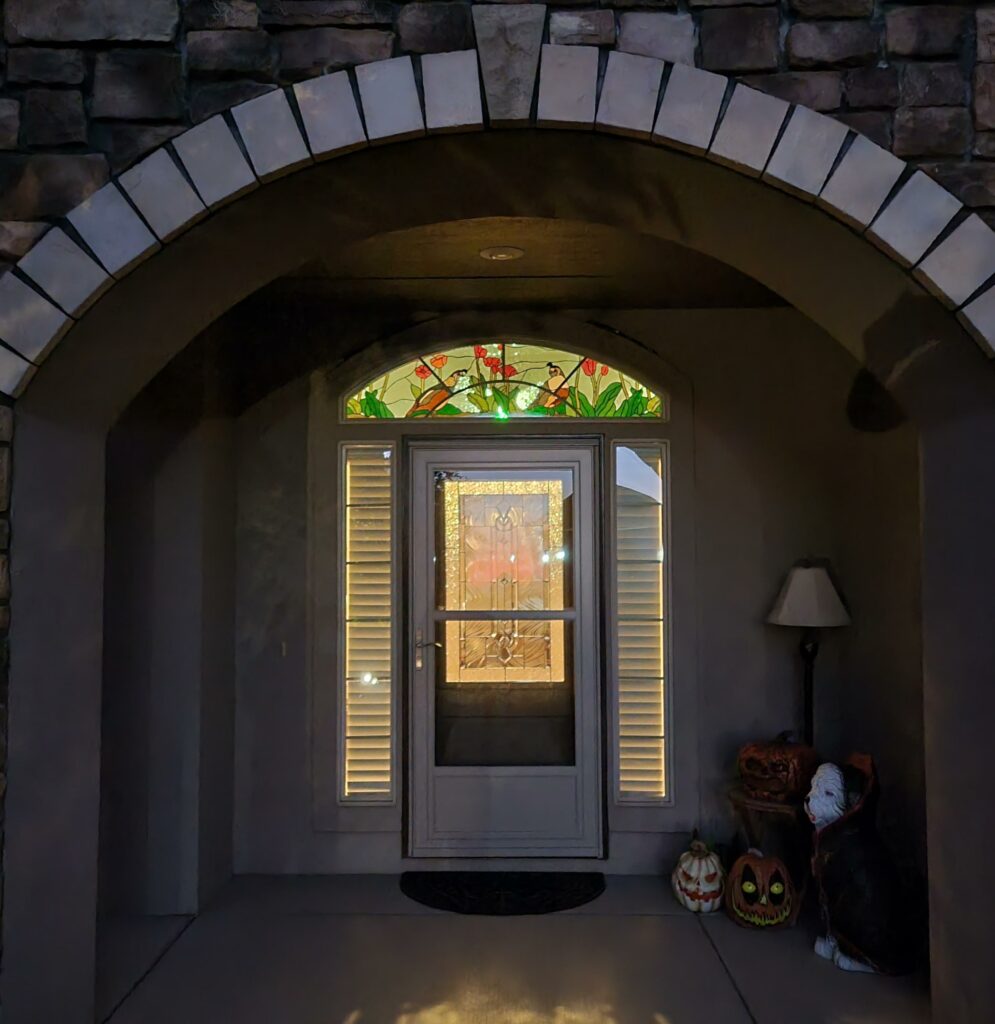 An arched stained glass transom window depicting two quail birds among red tulip flowers and green leaves, set above a front door with a clear beveled glass insert and flanked by sidelights, all within a stone arched entryway at night.