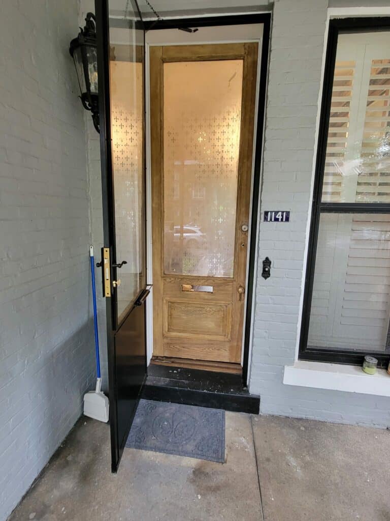 A wooden front entry door featuring an upper glass panel with a frosted fleur-de-lis pattern for privacy, seen behind a partially open black storm door. The house has gray brick siding.