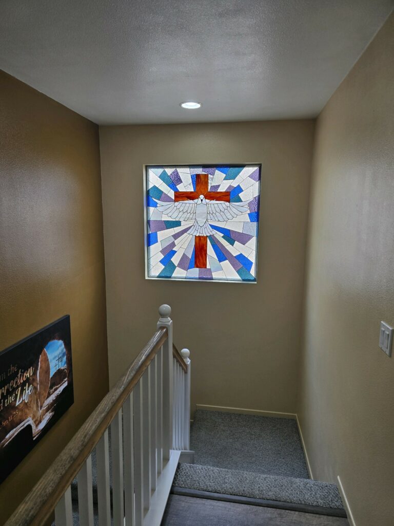 A square stained glass window depicting a white dove with outstretched wings centered over a reddish-brown cross. The background features radiating geometric panels in shades of blue, purple, and clear textured glass, creating a radiant effect.