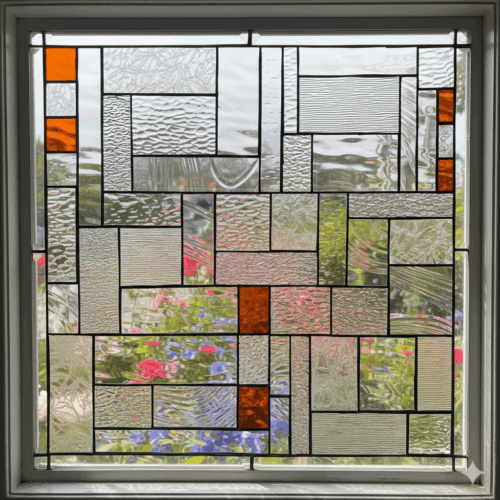 A square stained glass window with a modern, abstract geometric pattern. The design is a collage of many different clear, textured glass rectangles, accented with small squares of bright orange glass.
