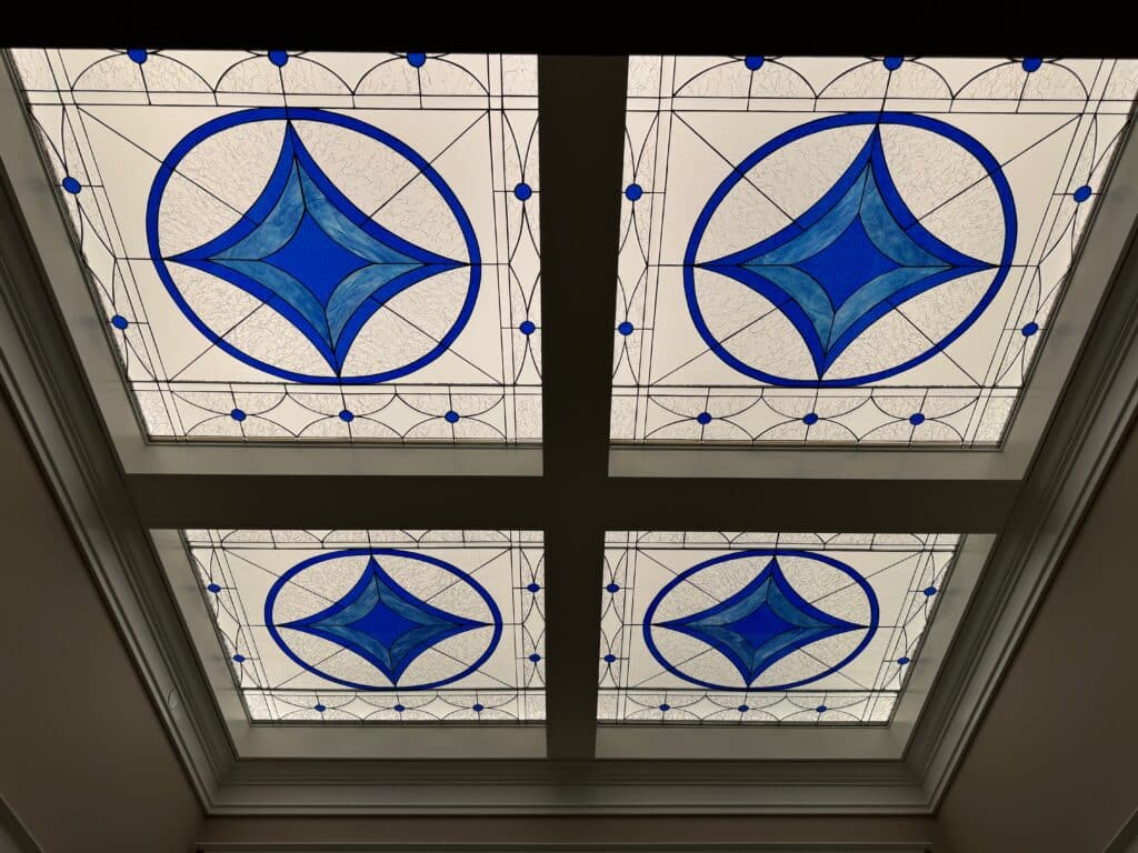 Four rectangular stained glass skylight panels featuring a central oval design with a vibrant blue diamond shape, surrounded by clear textured glass.