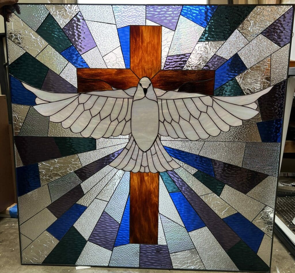 A square stained glass panel features a white dove with outstretched wings centered in front of a brown cross, surrounded by radiating beams of textured clear, blue, purple, and green glass.