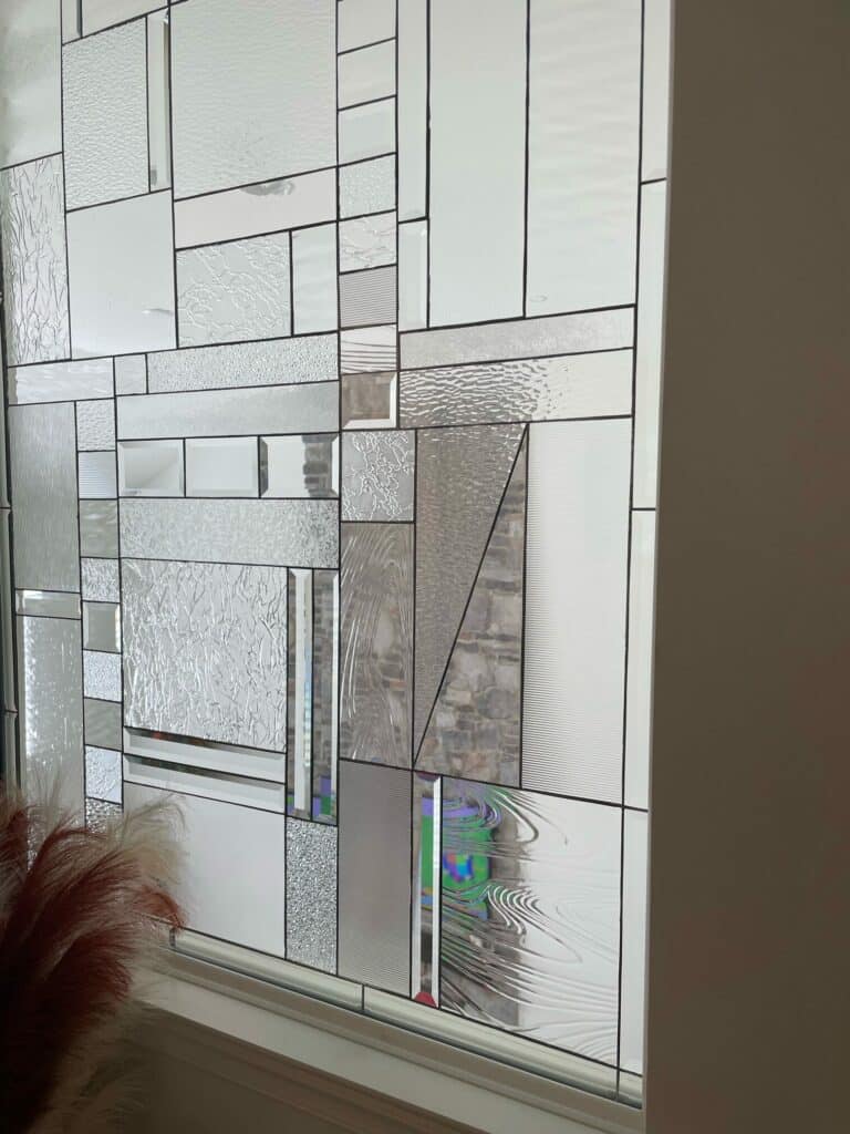 A rectangular, leaded stained glass window featuring an abstract geometric design composed of various clear textured, beveled, and mirrored glass pieces.