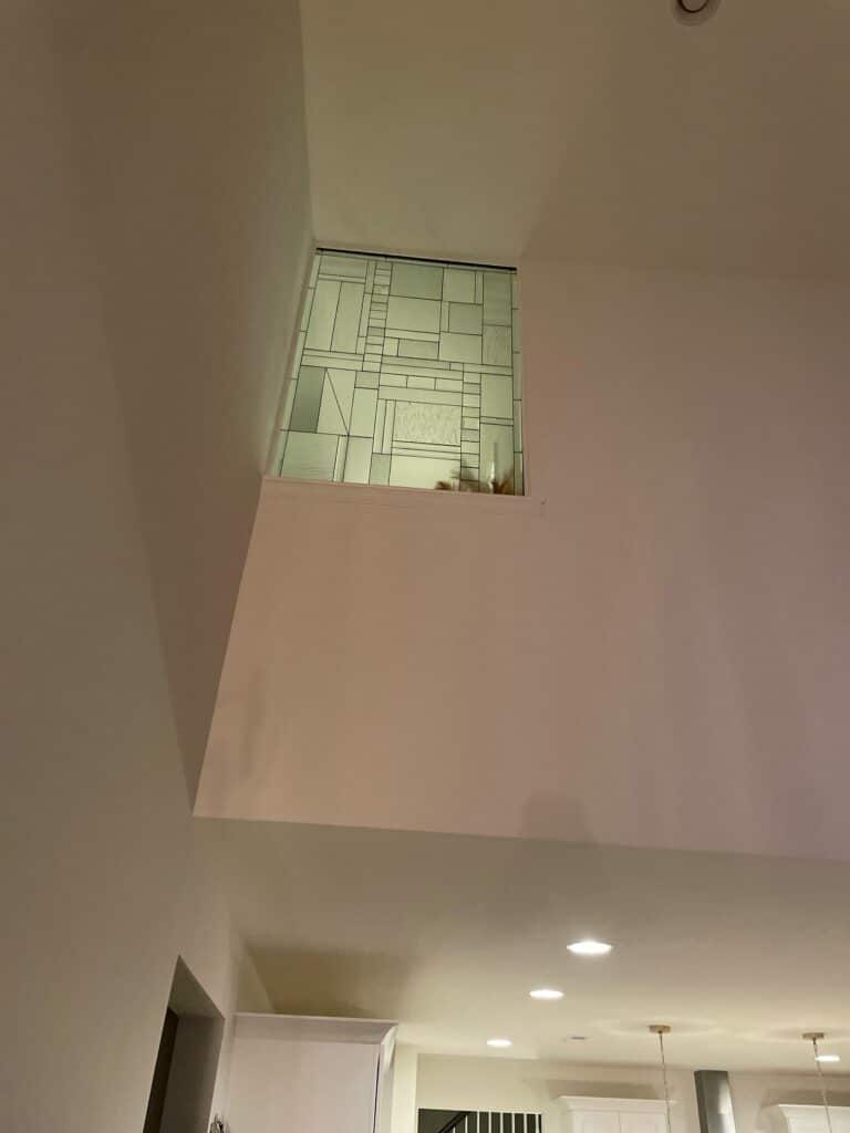 A rectangular, leaded stained glass window with a modern abstract geometric design, installed high in a white wall.