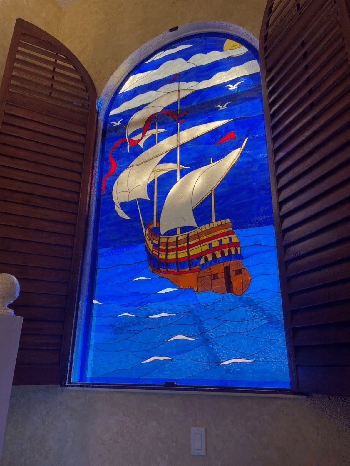 An arched stained glass window depicting a large, colorful sailing ship with white sails on a vibrant blue sea, under a blue sky with white clouds and flying birds. The window is framed by open dark wooden shutters.